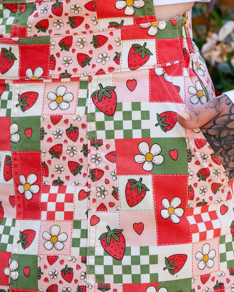 Super close up of a woman with tattoos wearing a strawberry picnic checkerboard patterned dress with a white long sleeve underneath. They have their hand tucked into the pinafore dress pocket showing the zip detailing too. 
