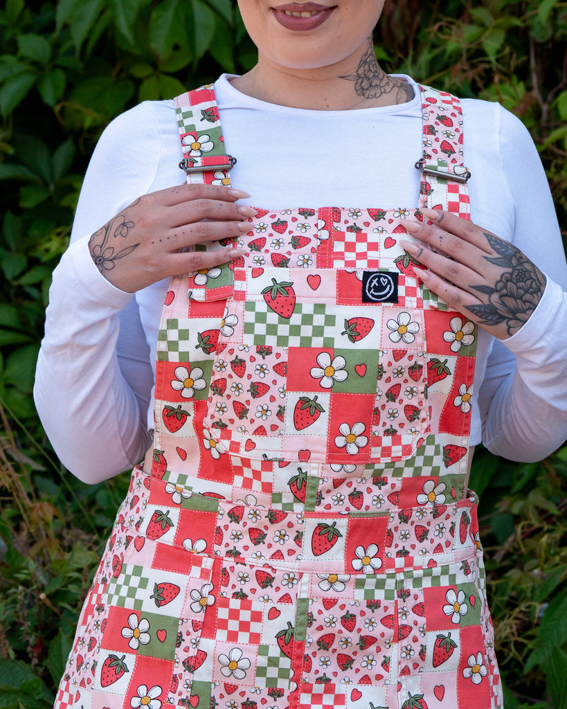Close up of a femme model with tattoos wearing the strawberry picnic pinafore dress with a white long sleeve top underneath. She is facing forward smiling whilst holding the adjustable straps. The pinafore print features blossoms, strawberries and checkerboard in sage green, pastel red, baby pink and white.