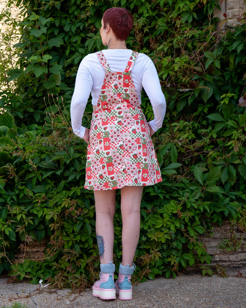 Alternative tattooed model with short reddy purple hair wearing the rebel romance strawberry picnic pinafore dress with a white long sleeve crop top and pink platform boots. She is facing away from the camera with her hands in her pockets outside in front of a lush green bush. The print on the pinafore is a sage green, pastel pink, pastel red and white colourblocks with strawberries and blossoms.