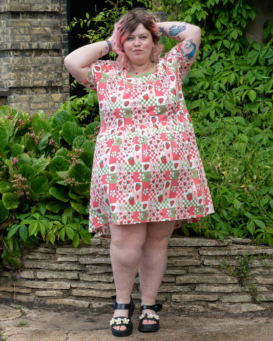 Non-binary model Sam wears the Rebel Romance Strawberry Picnic Stretch Smock Dress, which features a cheerful patchwork pattern of strawberries, daisies, gingham, and hearts, standing with hands behind head in front of a stone wall and greenery.