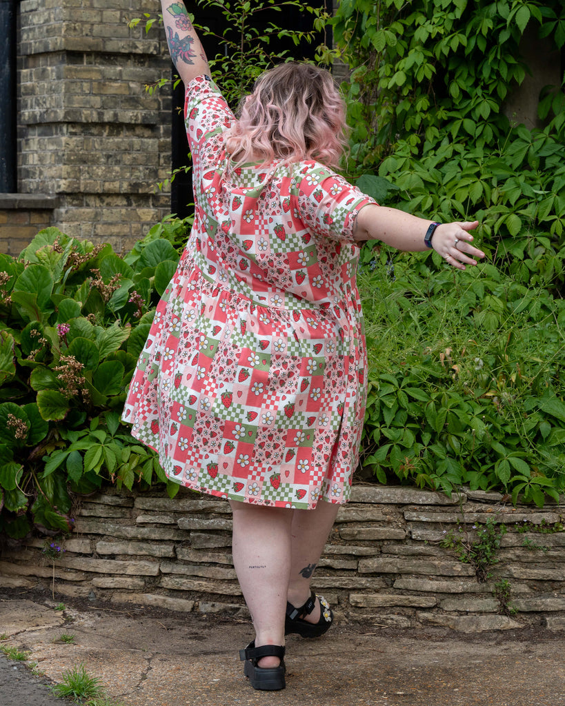 Back view of Sam twirling in the Rebel Romance Strawberry Picnic Stretch Smock Dress, showing the flared shape and all-over patchwork pattern of strawberries, daisies, hearts, and gingham.
