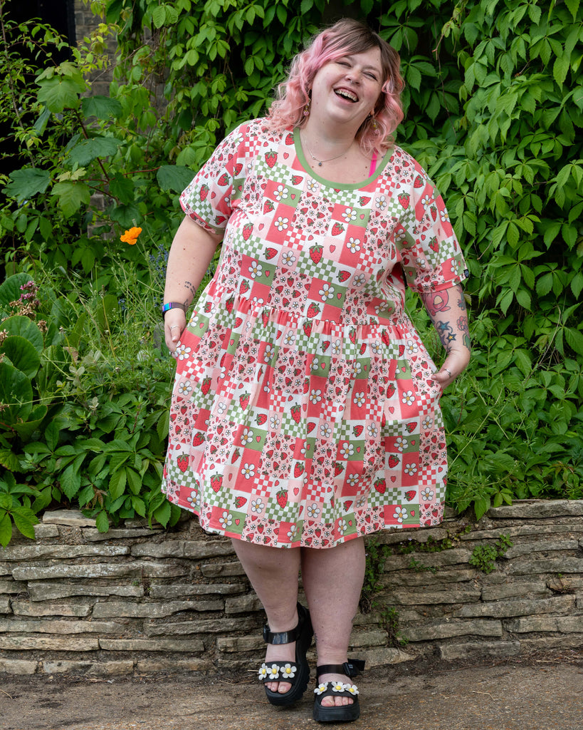 Sam poses joyfully in the Rebel Romance Strawberry Picnic Stretch Smock Dress, which is printed with a fun and nostalgic patchwork of red strawberries, white daisies, gingham squares, and pink hearts.