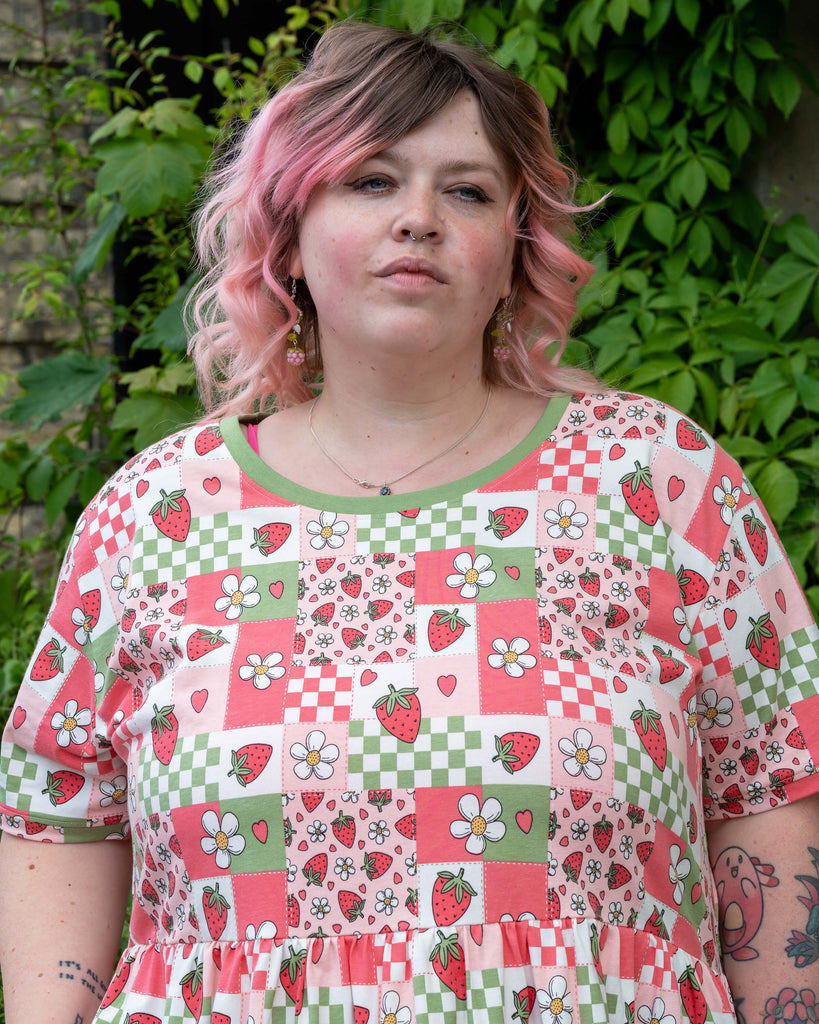 Close-up portrait of Sam wearing the Rebel Romance Strawberry Picnic Stretch Smock Dress, showing the neckline and the vibrant patchwork of strawberries, hearts, white daisies, and green checkerboard print.