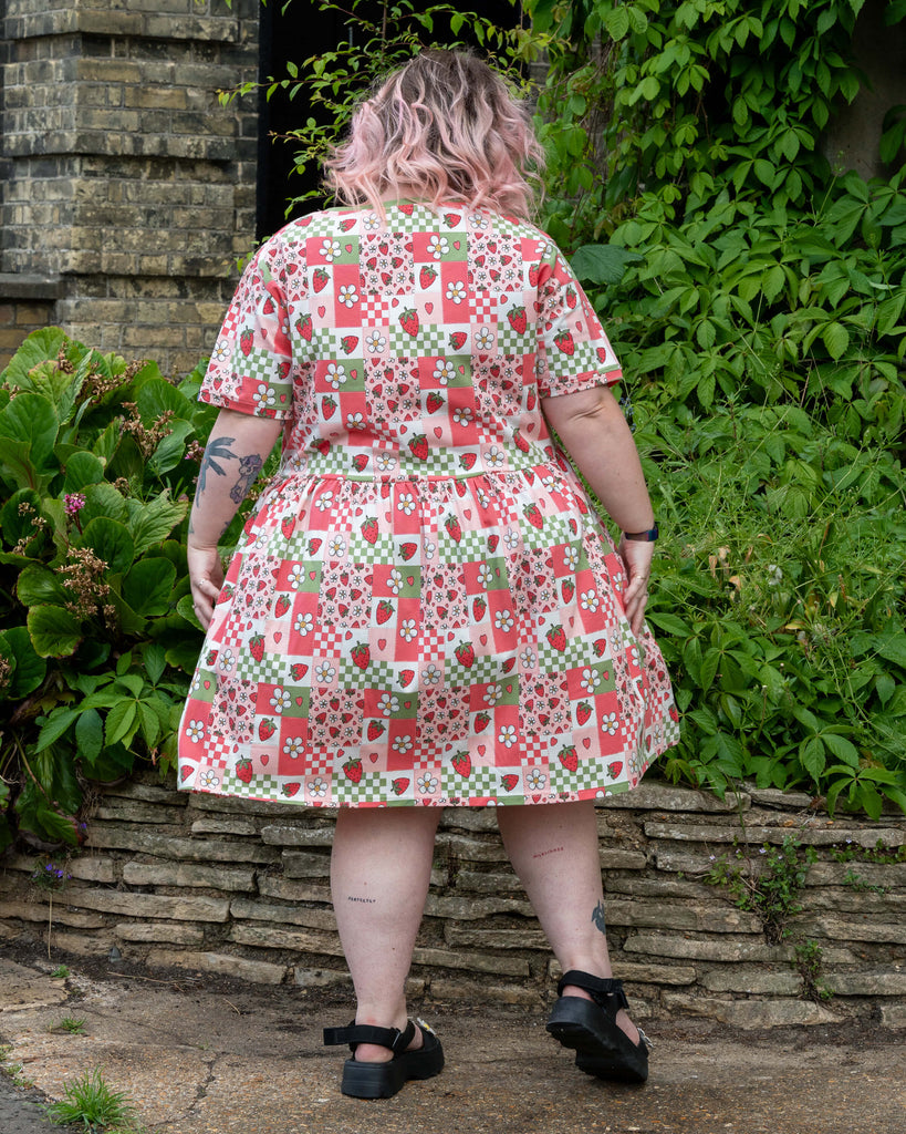 Back view of the Rebel Romance Strawberry Picnic Stretch Smock Dress worn by Sam, highlighting the gathered skirt and colourful patchwork pattern of strawberries, daisies, gingham, and hearts.