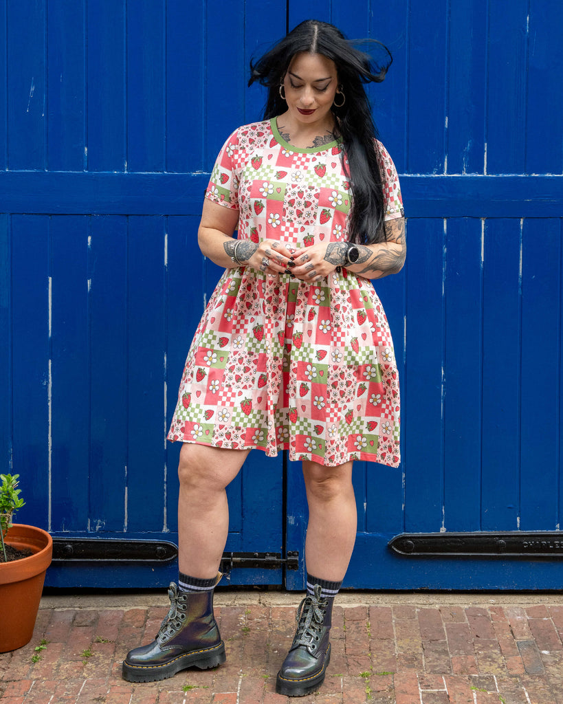 Carmen stands in front of a bold blue wooden door wearing the Rebel Romance Strawberry Picnic Stretch Smock Dress. The short-sleeve dress features a fun pink, green, and red patchwork print with strawberries, daisies, hearts, and checks. She styles it with iridescent lace-up boots and striped socks. Her long black hair is down and blowing gently in the breeze.