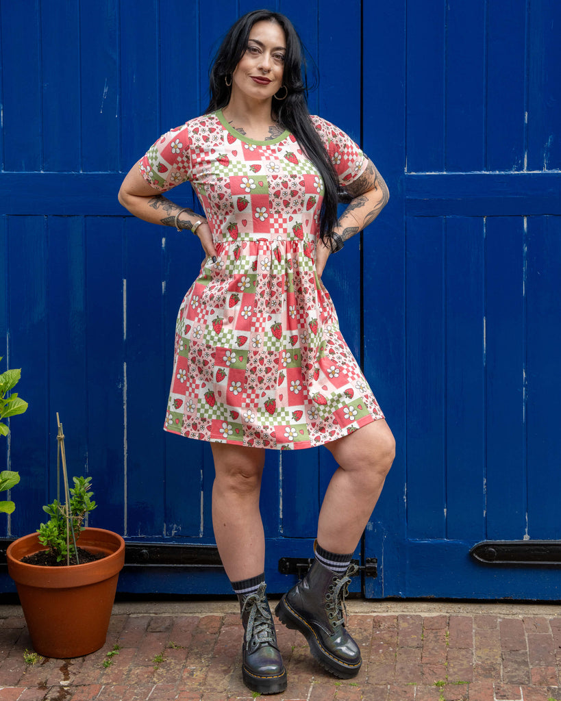 Carmen poses with hands on hips in the Rebel Romance Strawberry Picnic Stretch Smock Dress, standing confidently in front of a bright blue wooden backdrop. The short dress flares at the waist and is paired with shiny lace-up boots and stripey socks for a bold, alternative look.