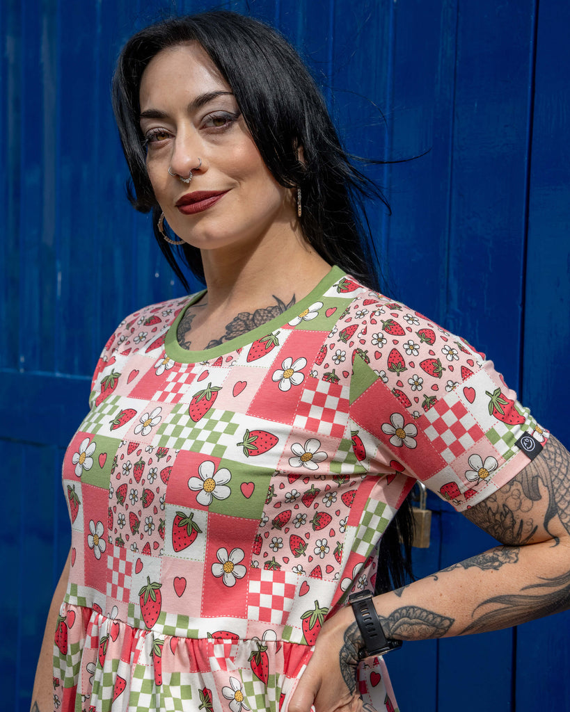 Close-up of Carmen wearing the Rebel Romance Strawberry Picnic Stretch Smock Dress, showing the cute patchwork print of strawberries, flowers, hearts, and checkerboard details. The green neckline trim contrasts with the soft pink and red tones. Her tattoos and hoop earrings are visible, and she smiles slightly in front of a vibrant blue door.