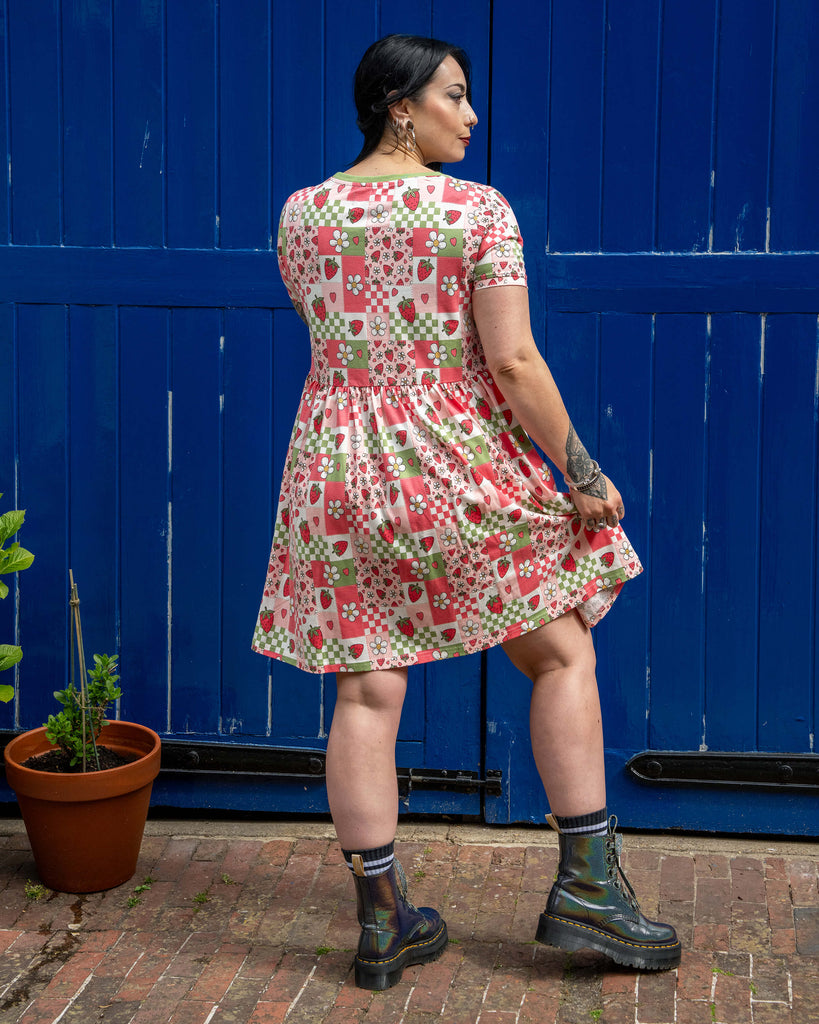 Back view of Carmen wearing the Rebel Romance Strawberry Picnic Stretch Smock Dress, showing the full print across the back. The dress has a soft gather at the waist and flows out in an A-line shape. She pairs it with iridescent boots and poses in front of a blue door with brick flooring.