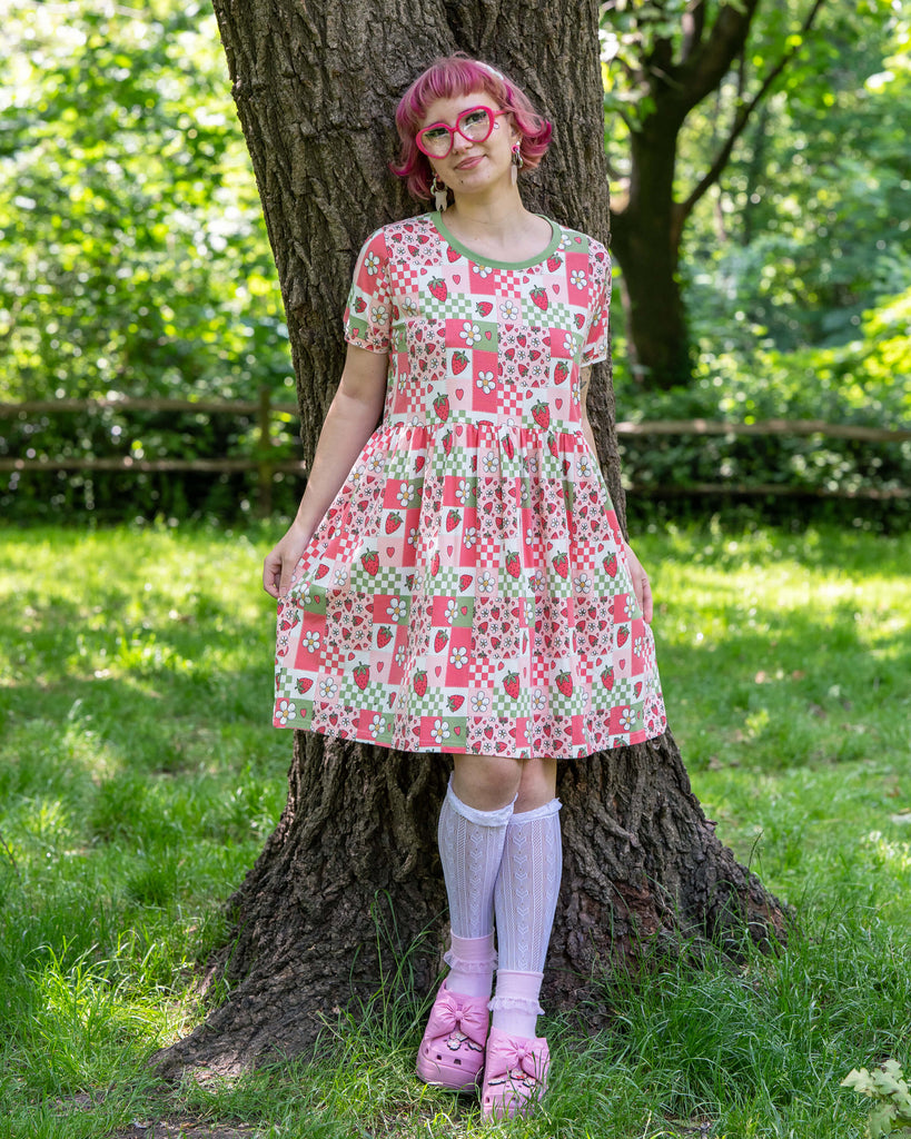 A model with bobbed pink hair, Jess, is wearing the rebel romance strawberry picnic oversized smock dress, white knee high socks, pink heeled crocs and pink accessories. The dress print is sage green, pastel red, baby pink and white repeating tiles of strawberries, checkerboard, flower blossoms and hearts, the neckline is a contrasting sage green colour. She is stood holding out the bottom of the dress and leaning back against a tree smiling outside in a park. 