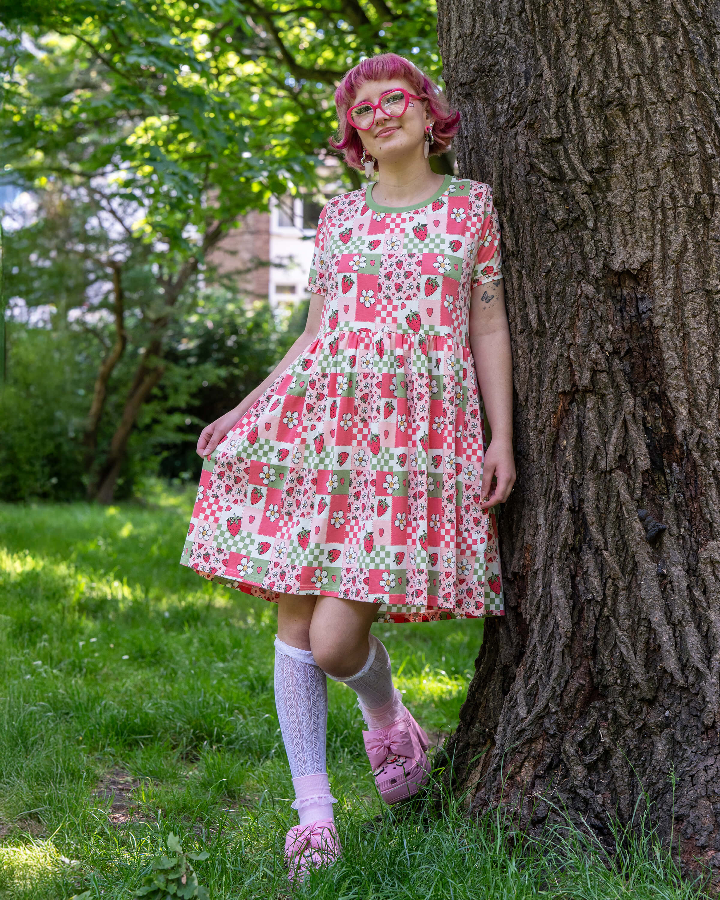 A model with bobbed pink hair, Jess, is wearing the rebel romance strawberry picnic oversized smock dress, white knee high socks, pink heeled crocs and pink accessories. The dress print is sage green, pastel red, baby pink and white repeating tiles of strawberries, checkerboard, flower blossoms and hearts, the neckline is a contrasting sage green colour. She is stood in a lush green park area smiling to camera leaning against a tree.