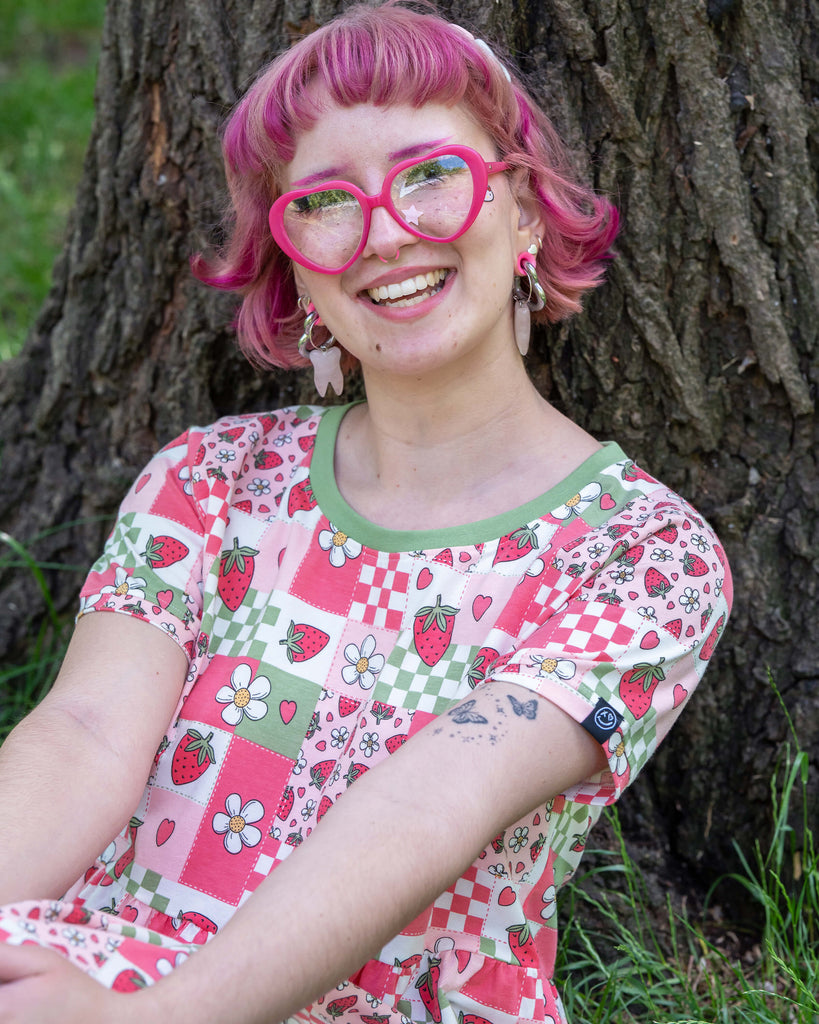 Close up of a model with bobbed pink hair, Jess, is wearing the rebel romance strawberry picnic oversized smock dress and pink accessories. The dress print is sage green, pastel red, baby pink and white repeating tiles of strawberries, checkerboard, flower blossoms and hearts, the neckline is a contrasting sage green colour. She is sat down in a park leaning against a tree laughing to camera.