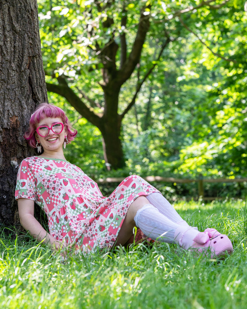 A model with bobbed pink hair, Jess, is wearing the rebel romance strawberry picnic oversized smock dress, white knee high socks, pink heeled crocs and pink accessories. The dress print is sage green, pastel red, baby pink and white repeating tiles of strawberries, checkerboard, flower blossoms and hearts, the neckline is a contrasting sage green colour. She is sat underneath a tree in a grassy park laughing looking off to the right.