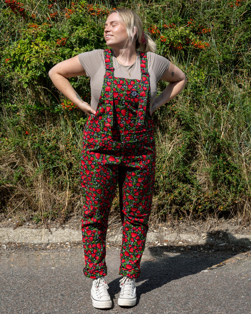 Harleigh wears Rebel Romance Strawberry Field stretch twill dungarees featuring a vibrant red strawberry print with green leaves on a black base. Styled with a beige top and chunky trainers for a bold alternative fashion look.