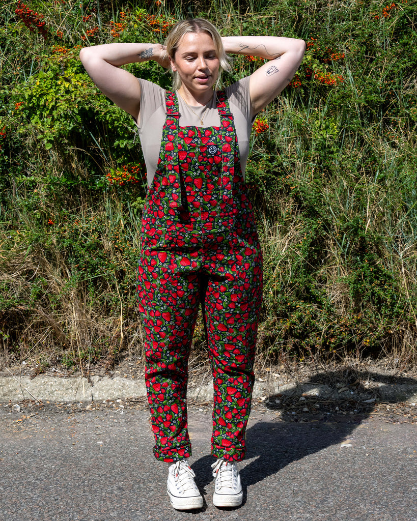 Harleigh wears Rebel Romance Strawberry Field stretch twill dungarees featuring a vibrant red strawberry print with green leaves on a black base. Styled with a beige top and chunky trainers for a bold alternative fashion look.