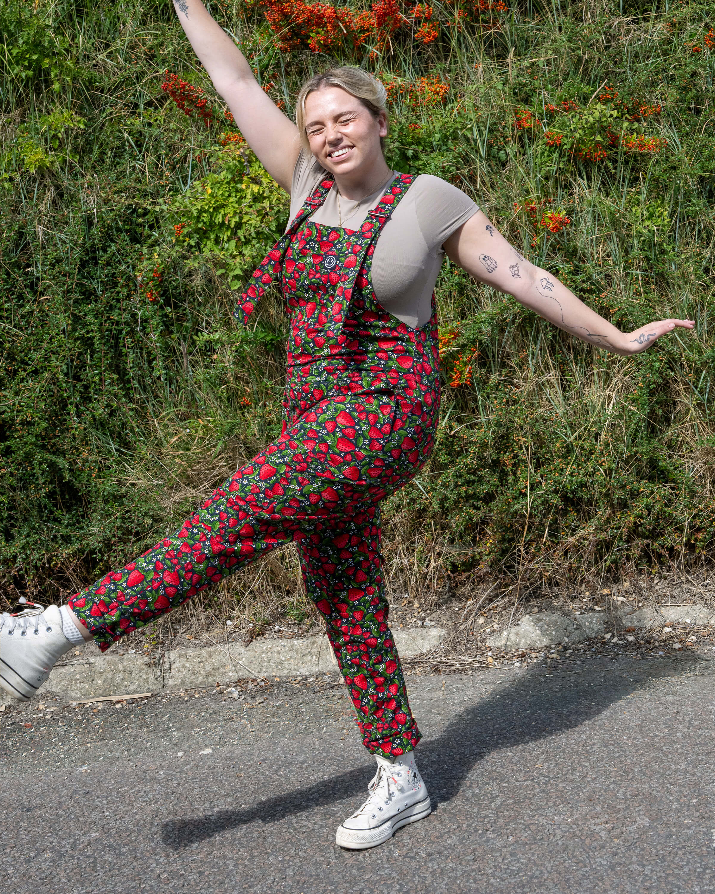 Harleigh poses in Rebel Romance Strawberry Field stretch twill dungarees with a bold red strawberry print. A fun dopamine dressing look for alternative and retro fashion lovers.