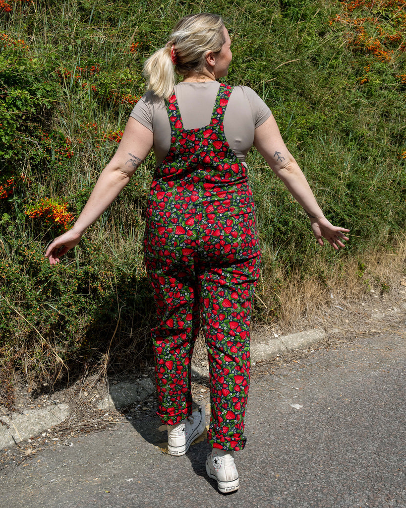 Back view of Harleigh in Rebel Romance Strawberry Field stretch twill dungarees, showing the all-over strawberry fruit print with adjustable straps and a fitted shape.