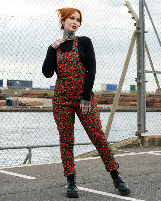 Erin wears Rebel Romance Strawberry Field stretch twill dungarees featuring a vibrant red strawberry print with green leaves on a black base. Styled with a black top and chunky boots for a bold alternative fashion look.