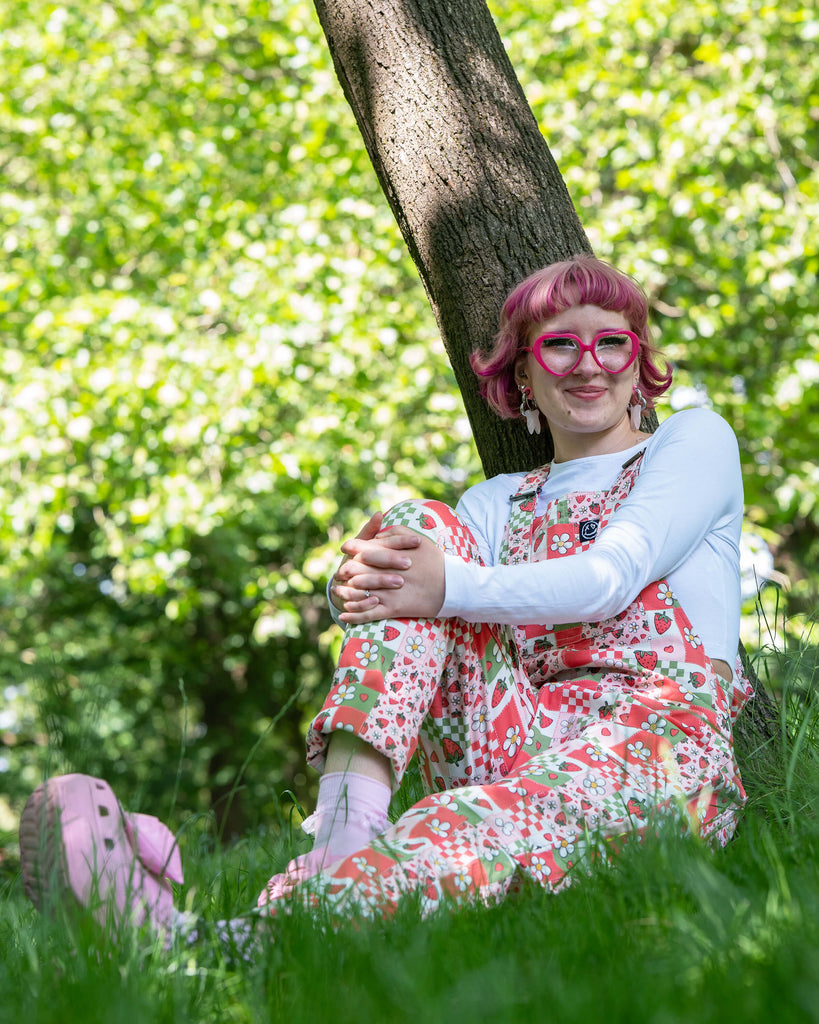 The strawberry picnic dungarees modelled on Jess, a pink haired model, with a white long sleeve top underneath and pink heeled crocs. The dungaree print is sage green, pastel red, baby pink and white repeating tiles of strawberries, checkerboard, flower blossoms and hearts. She is sat outside in a lush green park smiling with her hands resting on her knee.