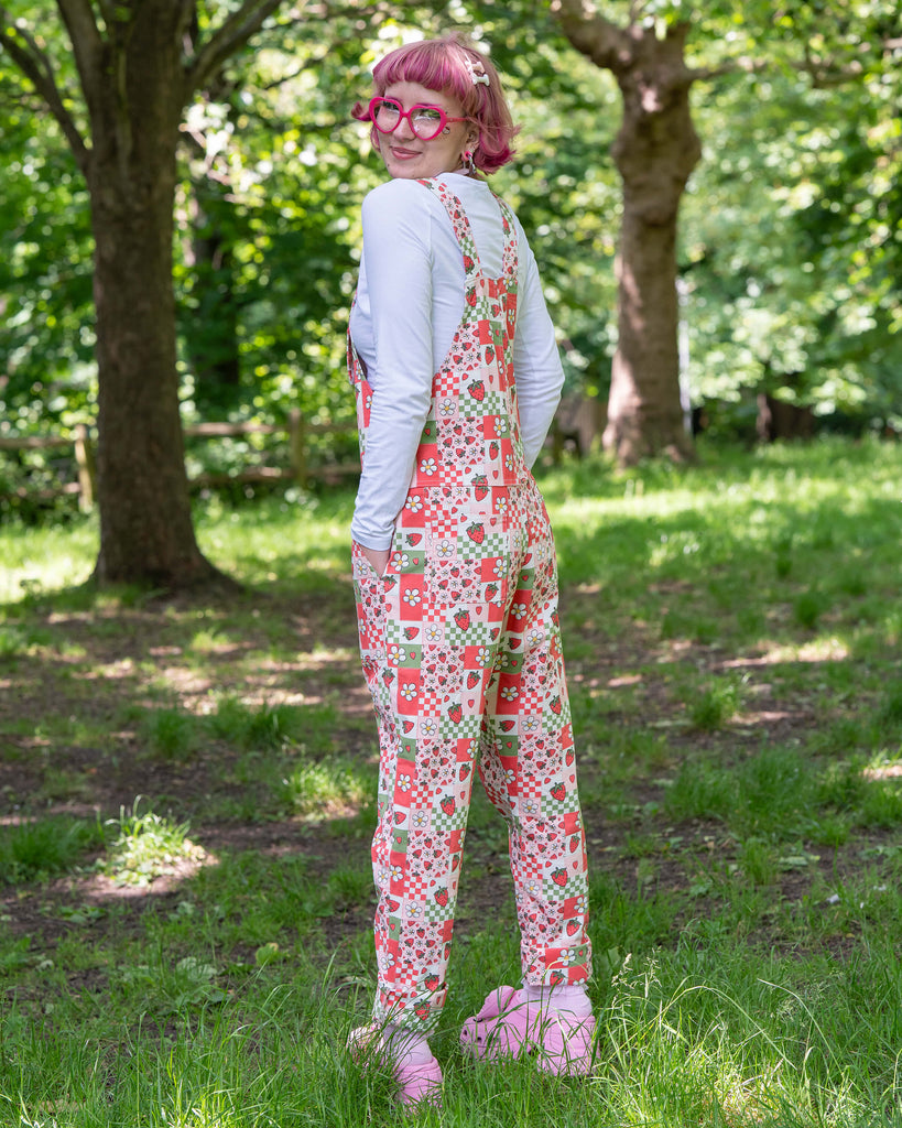 The strawberry picnic dungarees modelled on Jess, a pink haired model, with a white long sleeve top underneath. The dungaree print is sage green, pastel red, baby pink and white repeating tiles of strawberries, checkerboard, flower blossoms and hearts. She is stood outside in a lush green park facing away looking over her shoulder smiling with her hands resting in the dungaree pockets. 