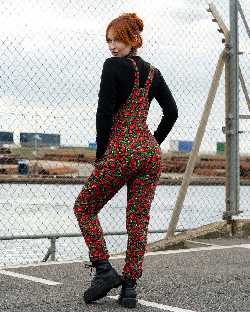Back view of Erin in Rebel Romance Strawberry Field stretch twill dungarees, showing the all-over strawberry fruit print with adjustable straps and a fitted shape.