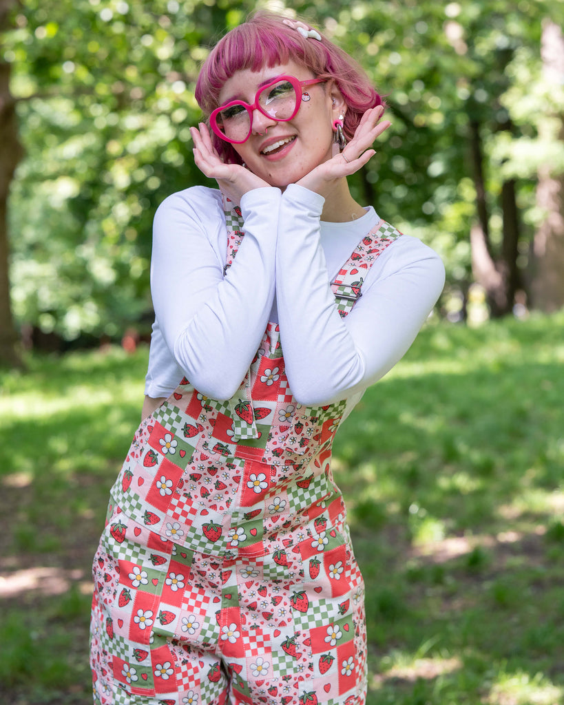 Close up of the strawberry picnic dungarees modelled on Jess, a pink haired model, with a white long sleeve top underneath. The dungaree print is sage green, pastel red, baby pink and white repeating tiles of strawberries, checkerboard, flower blossoms and hearts. She is stood outside in a lush green park smiling with her hands resting underneath her chin. 