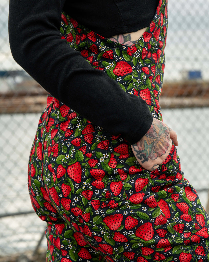 Close-up of the Strawberry Field dungarees by Rebel Romance, highlighting the juicy red strawberries, green leaves, and white blossoms against a black background.