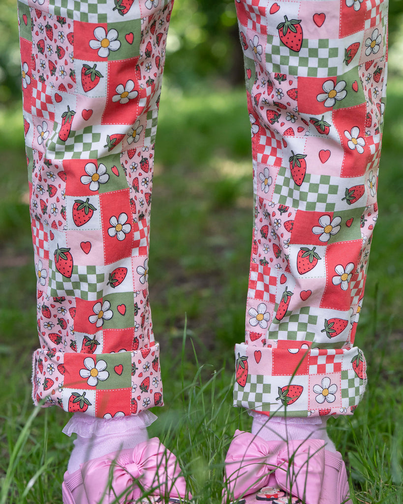 Close up of the strawberry picnic dungarees modelled on Jess with pink heeled crocs. The dungaree print is sage green, pastel red, baby pink and white repeating tiles of strawberries, checkerboard, flower blossoms and hearts. She is stood outside in a lush green park. 
