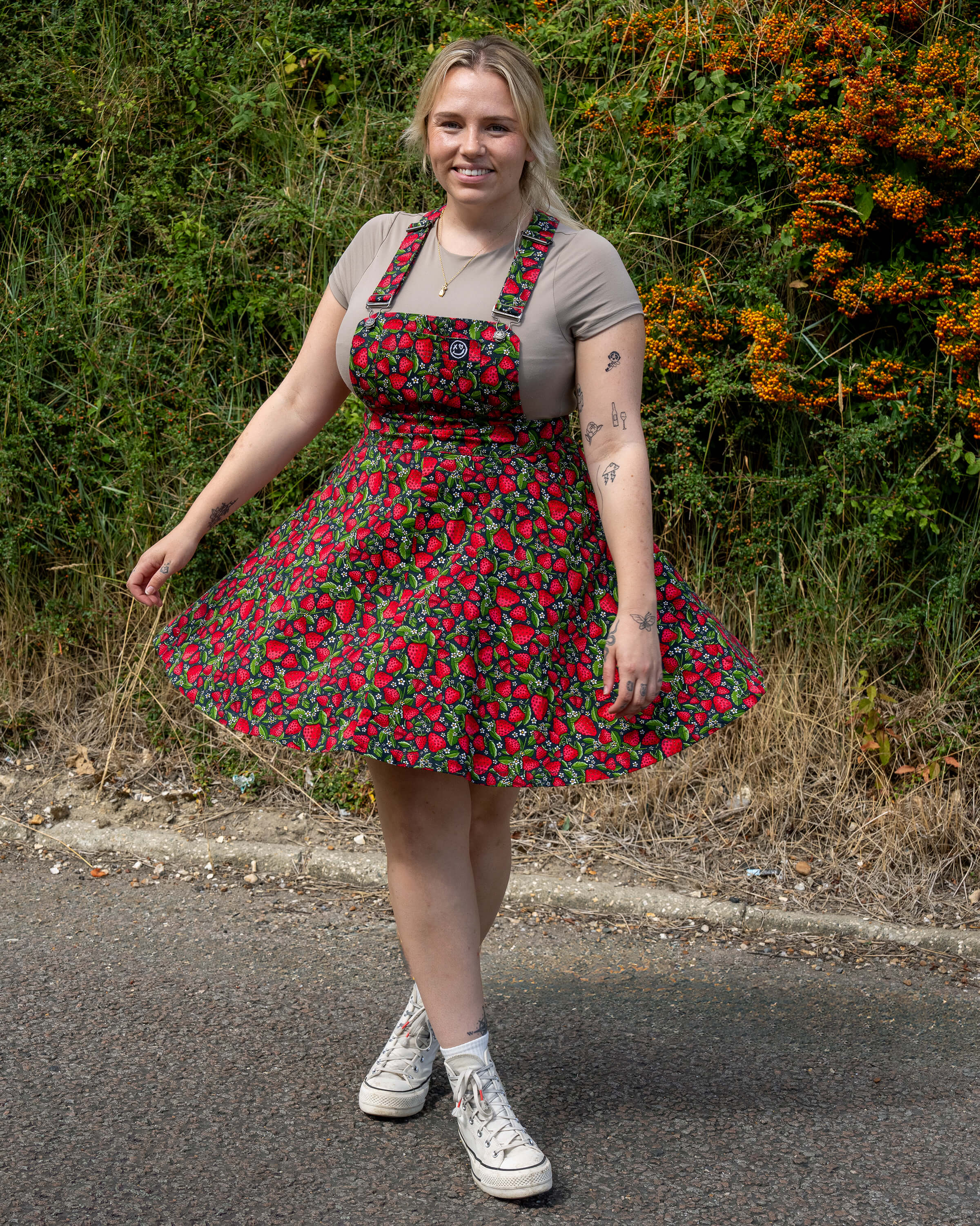 Model Harleigh wears the Rebel Romance Strawberry Field Flared Pinafore with a beige tee and Converse trainers, showing off the flared skirt and playful fruit pattern.