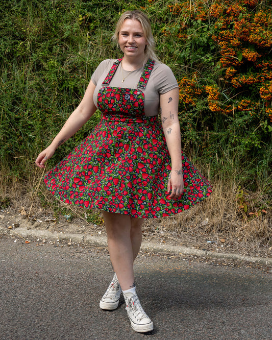 Model Harleigh wears the Rebel Romance Strawberry Field Flared Pinafore with a beige tee and Converse trainers, showing off the flared skirt and playful fruit pattern.