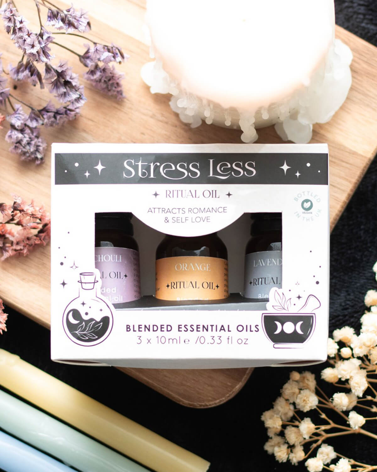  A box set of three Stress Less Ritual Blended Essential Oils (Patchouli, Lavender, and Orange) placed on a wooden tray with flowers and pastel-coloured candles, creating a calming, nature-inspired aesthetic.