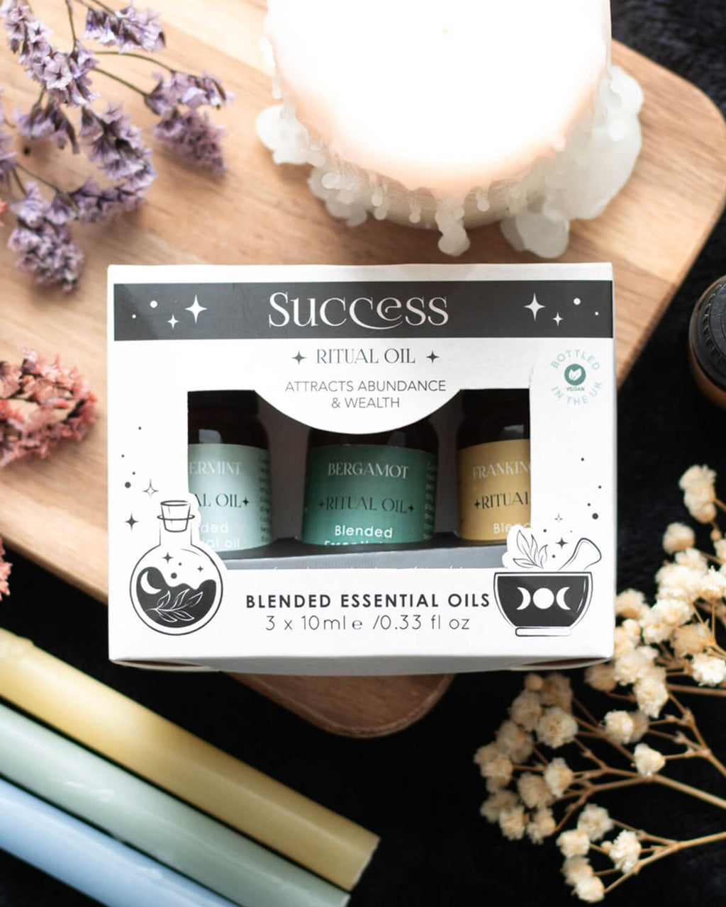  A box of ‘Success Ritual Oil’ essential oils with three visible 10ml bottles labeled Peppermint, Bergamot, and Frankincense. The packaging has a mystical design with stars, moons, and herbal illustrations, placed on a wooden board with dried flowers and candles.