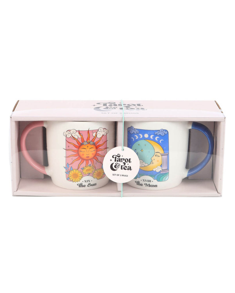A boxed set of two ceramic mugs featuring vibrant tarot-style designs of the Sun and Moon, with pink and blue handles visible through the packaging window. The label reads “Tarot & Tea” with “Set of 2 Mugs” on a round tag.