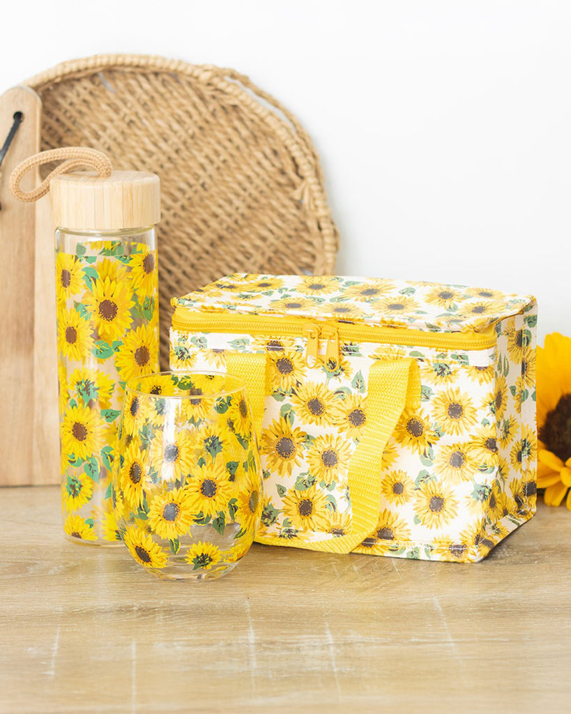 A sunflower-themed picnic set from Rebel Romance, a small UK business. The glass water bottle, insulated lunch bag, and tumbler all feature a vibrant sunflower print. The bottle has a bamboo lid with a cord loop, making it perfect for carrying on the go. A cheerful summer aesthetic aimed at millennials who love alternative style and retro vibes.