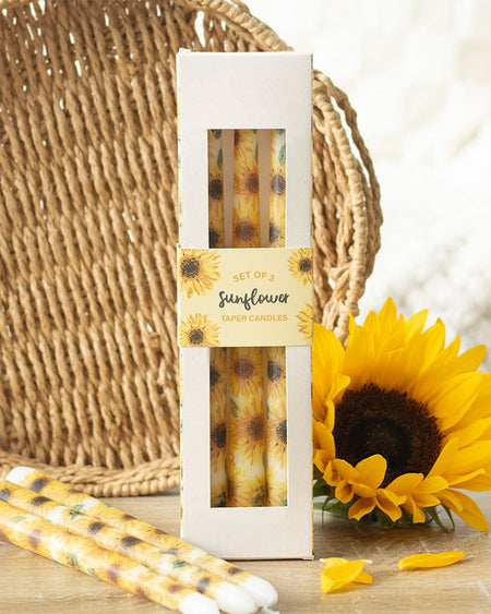 The sunflower taper candles in their original packaging, displayed against a woven basket background with a real sunflower beside them. The sunflower-themed box matches the candle design, creating a cohesive look. A fun and unique home accessory from Rebel Romance’s summer-inspired range.