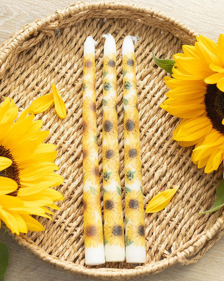 Three sunflower taper candles displayed in a woven basket alongside real sunflowers. The candles have vibrant sunflower prints on a cream-coloured wax base. Sunflower petals are scattered around for a summery aesthetic. A bright and cheerful addition to any alternative home, sold by Rebel Romance.