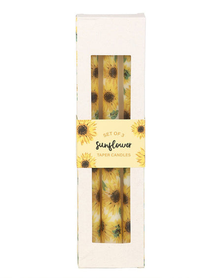 Close-up of the sunflower taper candles in their packaging, showing the bright yellow sunflower design with green leaves. The clear window in the box highlights the detailed print. The packaging has a sunflower border and a yellow label reading "Set of 3 Sunflower Taper Candles." A perfect gift for sunflower lovers, available at Rebel Romance.