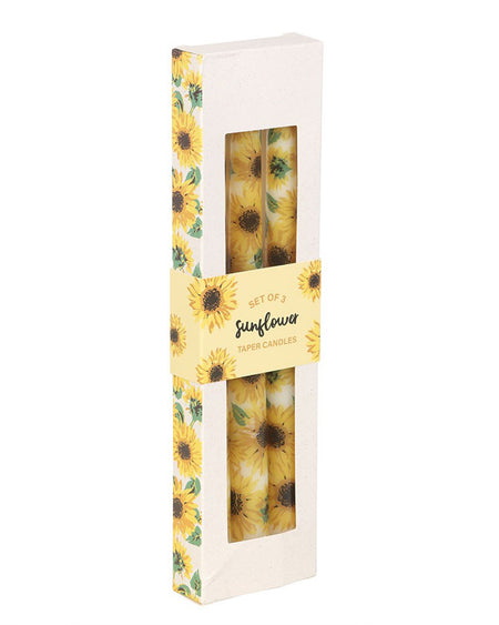 A set of 3 sunflower taper candles in a rectangular box with sunflower-themed packaging. The candles are bright yellow with realistic sunflower prints, visible through a clear window. The packaging has a sunflower pattern on the sides and a yellow label that reads "Set of 3 Sunflower Taper Candles." A cheerful home decor piece from Rebel Romance's alternative product range.
