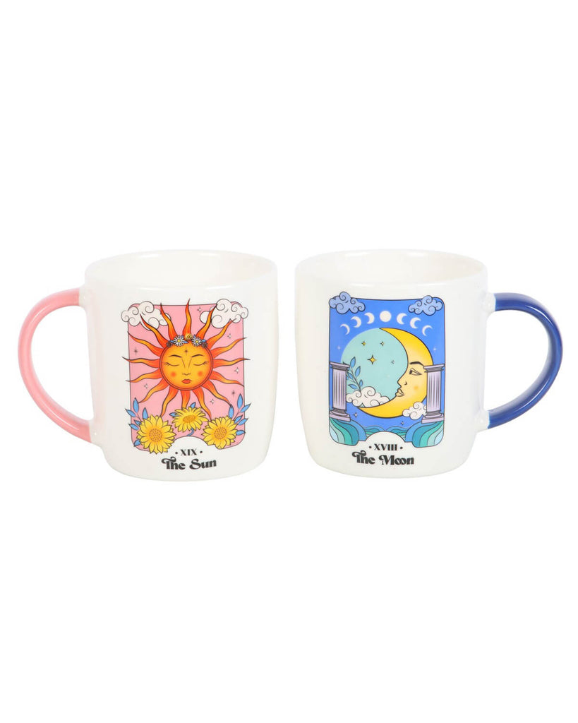 Two ceramic mugs side by side, one with a bright sun tarot design and pink handle, the other with a moon tarot design and blue handle, both featuring colourful celestial illustrations with clouds, stars, and floral motifs.