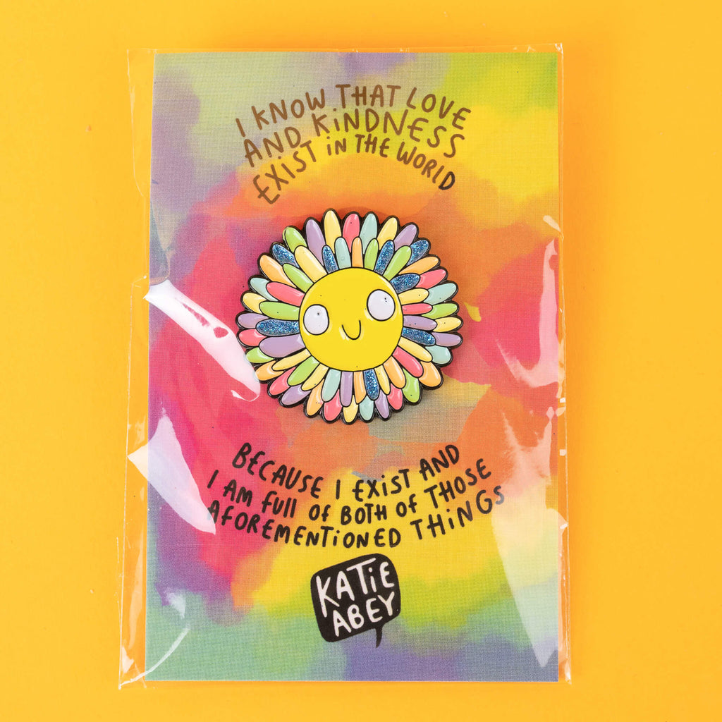 Katie Abey's enamel pin of a cheerful sun with rainbow petal rays, packaged on a vibrant backing card reading: 