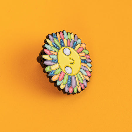 Close-up of Katie Abey's smiling sun enamel pin with glitter accents on its colourful rainbow rays. A bold and quirky accessory full of kindness and fun, perfect for adding a splash of joy to any outfit or bag.