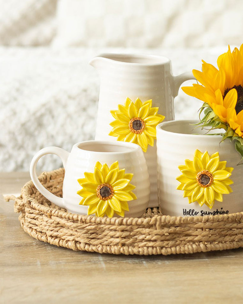 Cream ceramic mugs and a jug from Rebel Romance, each decorated with a bright 3D sunflower, displayed on a woven tray with fresh sunflowers. A warm, rustic homeware style perfect for adding cheerful charm to your kitchen.