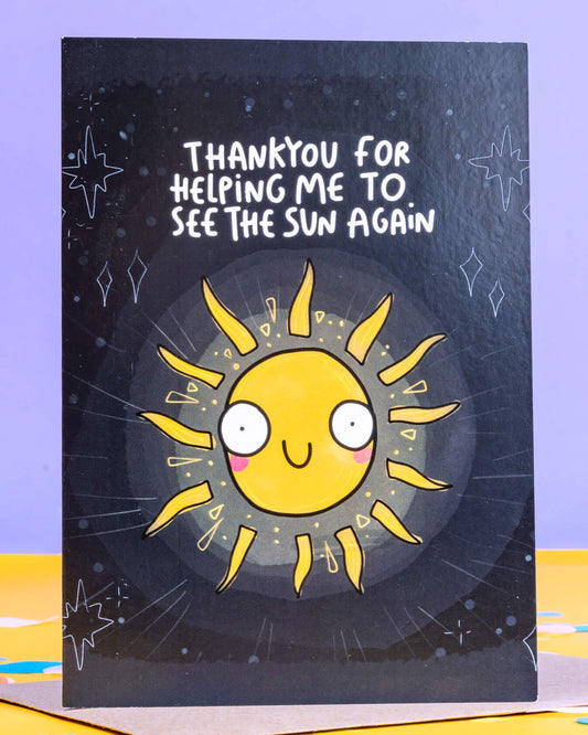 A6 greeting card from Rebel Romance featuring a bright, smiling cartoon sun with a cute face and rays against a dark sky. Surrounded by hand-drawn stars and sparkles, the card says “Thank you for helping me to see the sun again” in playful white text. A thoughtful and quirky illustrated card by Katie Abey, perfect for expressing gratitude. Designed in the UK by a small alternative brand celebrating unique style.