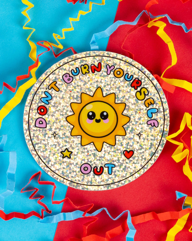 Round holographic glitter sticker featuring a smiling sun illustration and the text “Don’t Burn Yourself Out” in colourful bubble letters. The sticker is placed on a bright background of blue and red paper with colourful confetti strips. Designed by Innabox for Rebel Romance, a small UK brand celebrating alternative fashion and self-care.