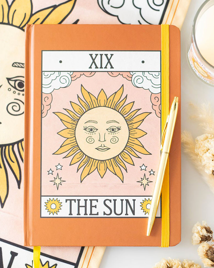 Flat lay of Rebel Romance’s The Sun Vintage Tarot A5 Lined Notebook featuring a vintage sun tarot design in yellow and pink on a burnt orange cover. Styled with a gold pen, floral elements and matching cloth. A dreamy and alternative gift idea from a UK indie brand that champions unique, artistic and witchy stationery for millennials.