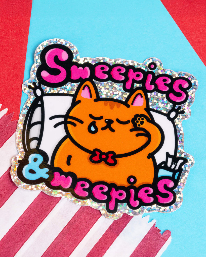 A holographic glitter vinyl sticker featuring a cute orange cat with a tear on its cheek, snuggled up on a pillow. The design reads “Sweepies & Weepies” in bold pink and black bubble letters, with shiny sparkles around the border. A fun and emotional sticker created by Innabox, available at Rebel Romance, a UK brand celebrating alternative style.