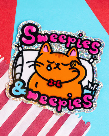 A holographic glitter vinyl sticker featuring a cute orange cat with a tear on its cheek, snuggled up on a pillow. The design reads “Sweepies & Weepies” in bold pink and black bubble letters, with shiny sparkles around the border. A fun and emotional sticker created by Innabox, available at Rebel Romance, a UK brand celebrating alternative style.