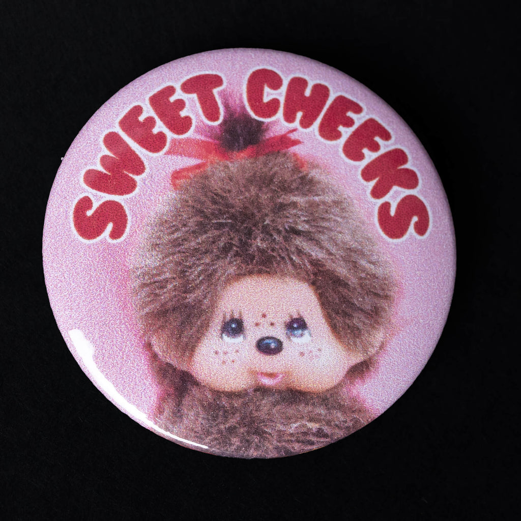 A 38mm high-gloss pin badge featuring a retro-style monkey with fluffy fur and a cheeky expression. The text “Sweet Cheeks” is written in bold red letters on a pink background. Designed by Bite Your Granny.