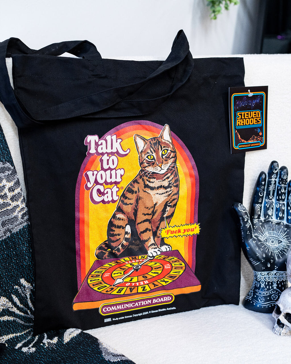 A black tote bag featuring a retro-inspired Steven Rhodes design titled “Talk to Your Cat.” The graphic shows a tabby cat sitting on a colourful communication board, with humorous text and a vintage aesthetic.