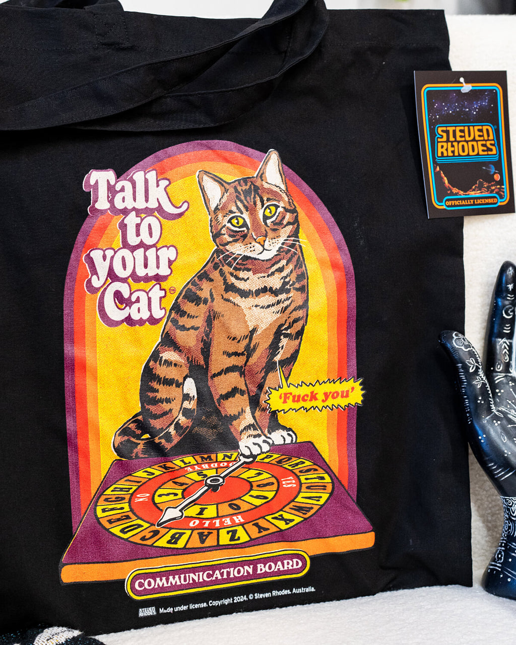 A black tote bag featuring a retro-inspired Steven Rhodes design titled “Talk to Your Cat.” The graphic shows a tabby cat sitting on a colourful communication board, with humorous text and a vintage aesthetic.