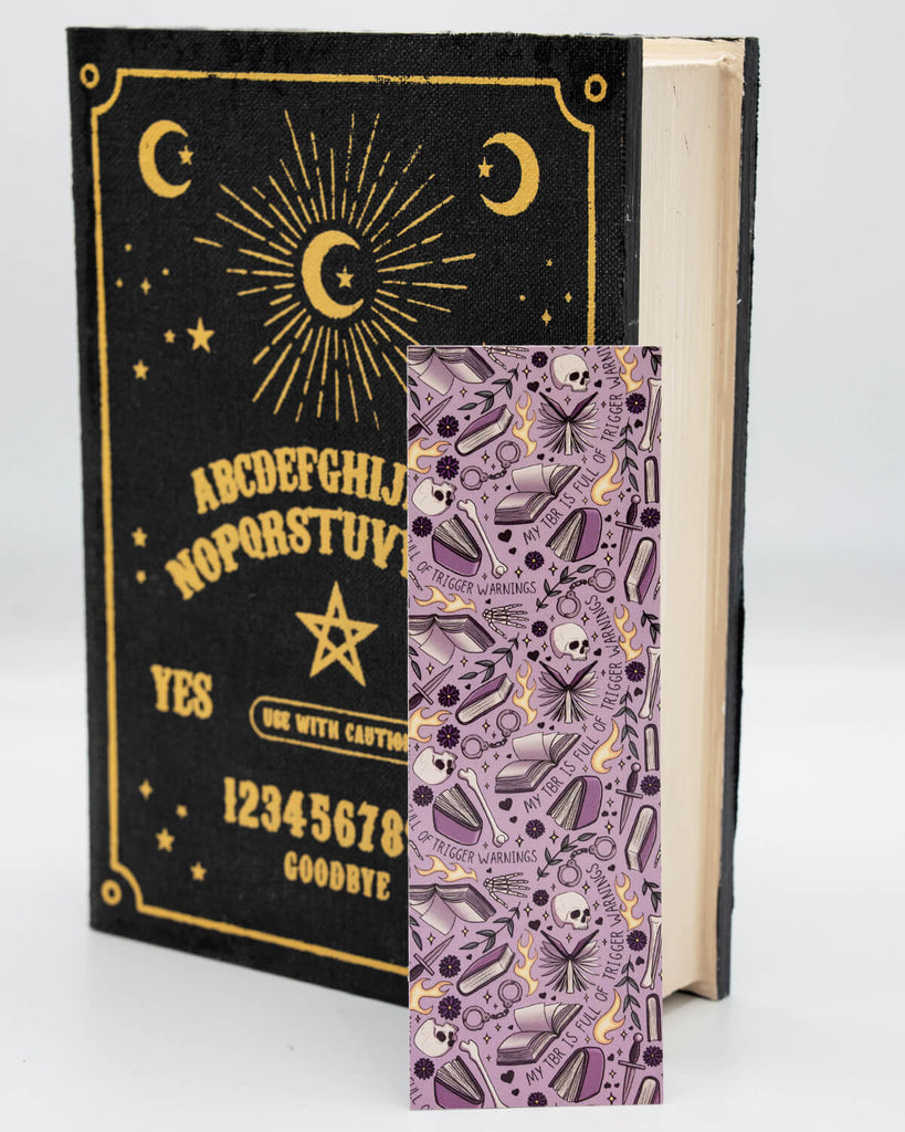 Purple gothic bookmark from Rebel Romance featuring a repeating print with skulls, flames, handcuffs, and the phrase “My TBR is full of trigger warnings.” Displayed inside a spooky black and gold book, this edgy accessory is perfect for dark romance readers and fans of taboo fiction.